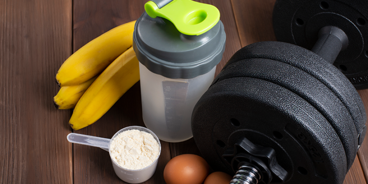 A scoop of protein with shaker and dumbbells with banana in the background.